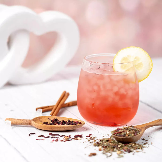A glass of iced Organic Sugar Crush Tea with a slice of lemon, surrounded by wooden spoons of loose leaf tea, hibiscus petals, and cinnamon sticks, with a soft heart-shaped decoration in the background.