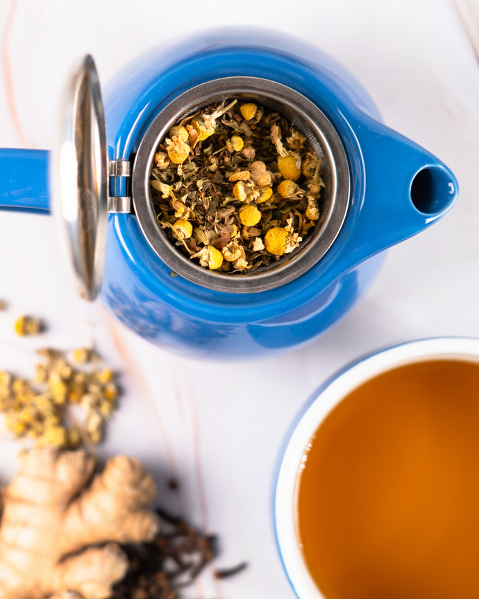 A close up image of Throat clarity tea herbs in a tea pot with a white mug of throat clarity tea besides it. 