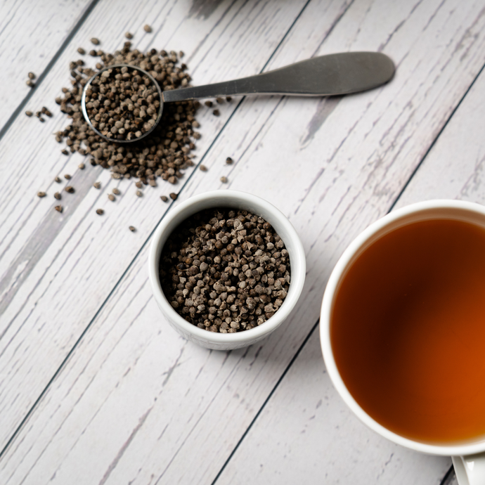 Vitex berries in a small white bowl and metal scoop beside a cup of brewed Organic Vitex Berry tea on a light wooden surface.