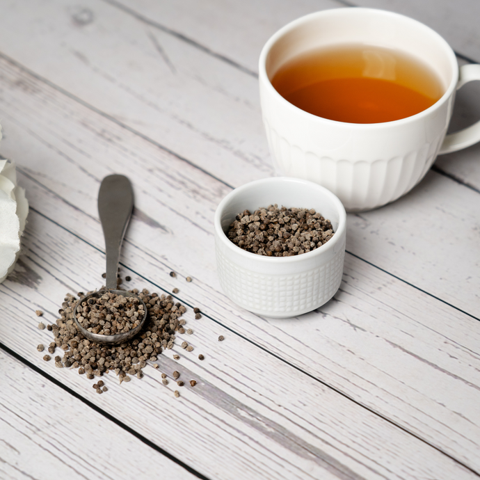 Vitex berries in a small white bowl and metal scoop beside a cup of brewed herbal tea on a light wood table.