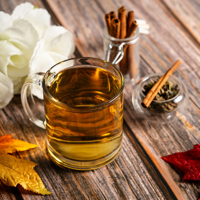 A glass mug of brewed Organic White Chai Tea on a rustic wooden table with cinnamon sticks, loose tea leaves, and autumn leaves beside white flowers.