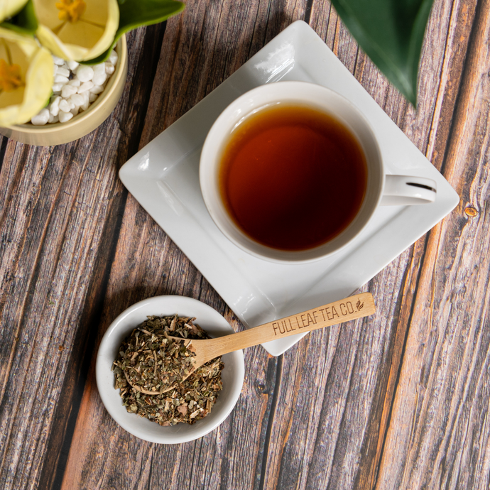 A cup of brewed Organic Wild Lettuce Tea on a white saucer beside a small bowl of loose leaf tea and a wooden spoon engraved with Full Leaf Tea Co., on a rustic wood surface with green plants nearby.